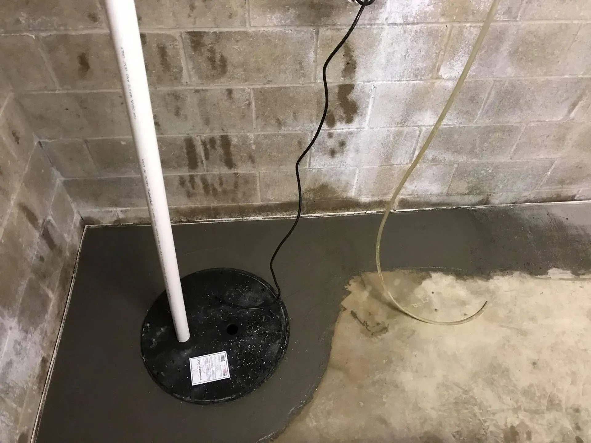 Basement utility area with sump pump lid, white pipe, cords, and wet concrete floor against cinderblock wall