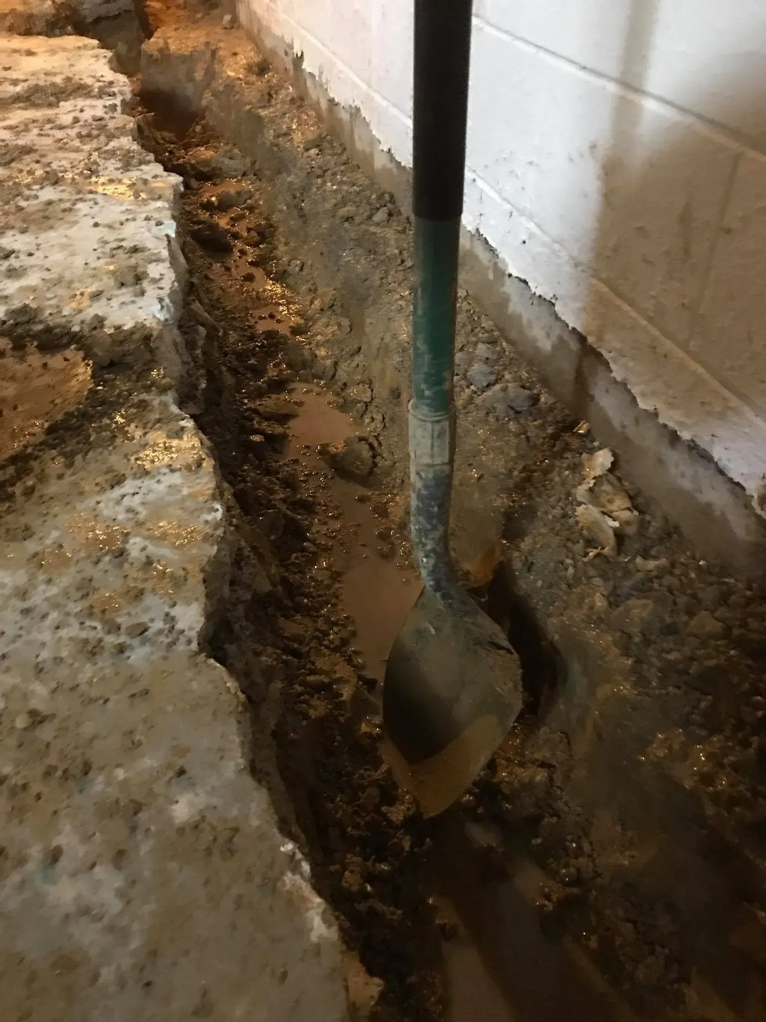 Shovel digging a narrow trench beside a cinder block wall along a concrete edge.