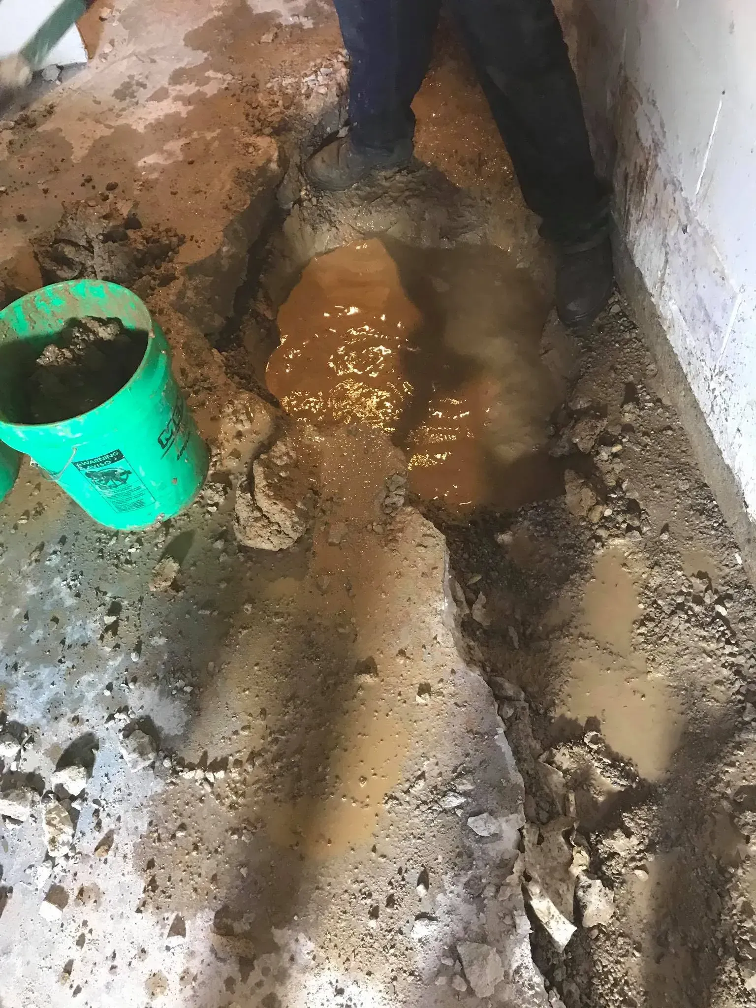 Fresh mud and standing water in a narrow trench beside a wall, with a green bucket nearby.