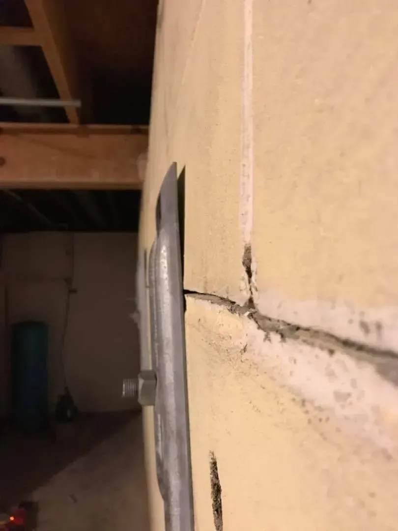Close-up of a cracked beige wall with a metal fixture in a dim basement ceiling area