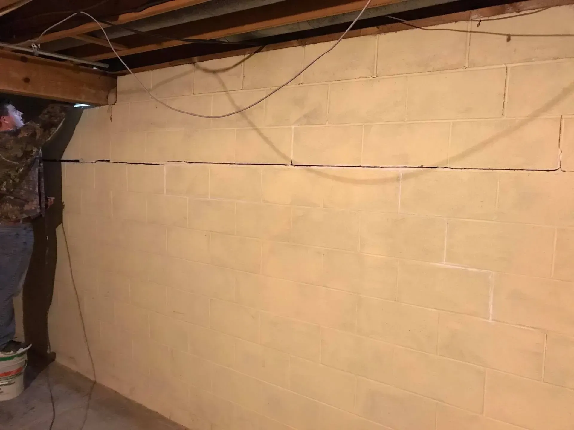 Basement cinderblock wall under exposed ceiling beams, partially painted beige with hanging wires.