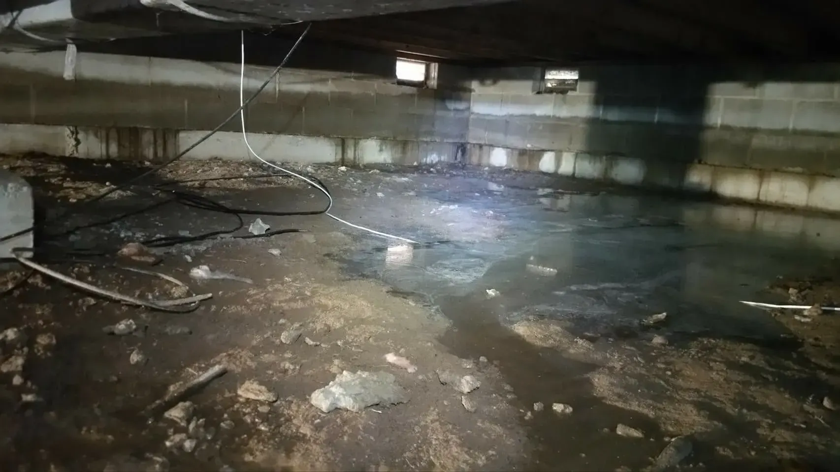 Dark flooded basement with standing water, debris, and exposed pipes under a low concrete ceiling