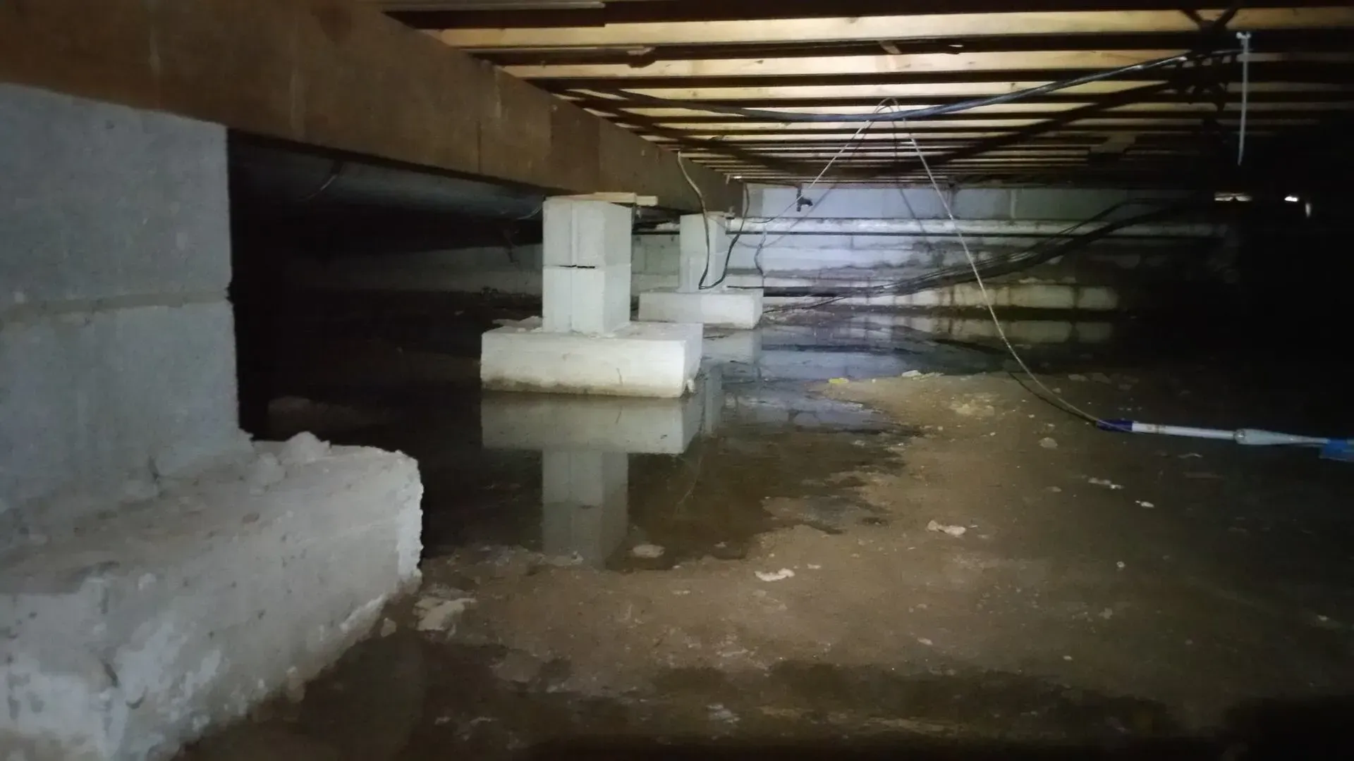 Flooded crawlspace under a wooden floor with support posts and standing water reflecting beams