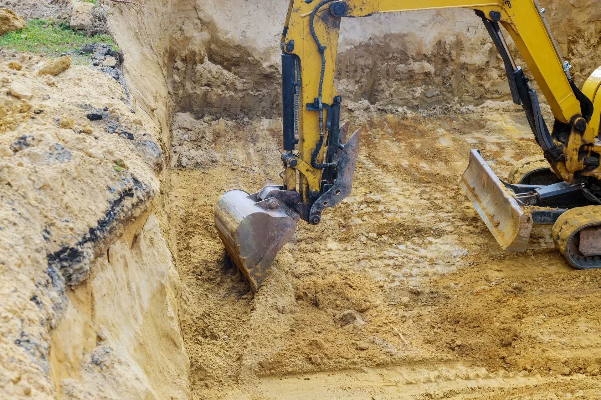 Excavator digging sandy soil at a construction site, with a rocky trench wall nearby