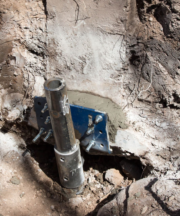 Metal pipe and blue bracket embedded in concrete patch on rough ground, with a hole in the dirt.