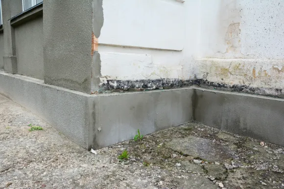 Corner of a white building with a damaged gray foundation and gravel ground around it