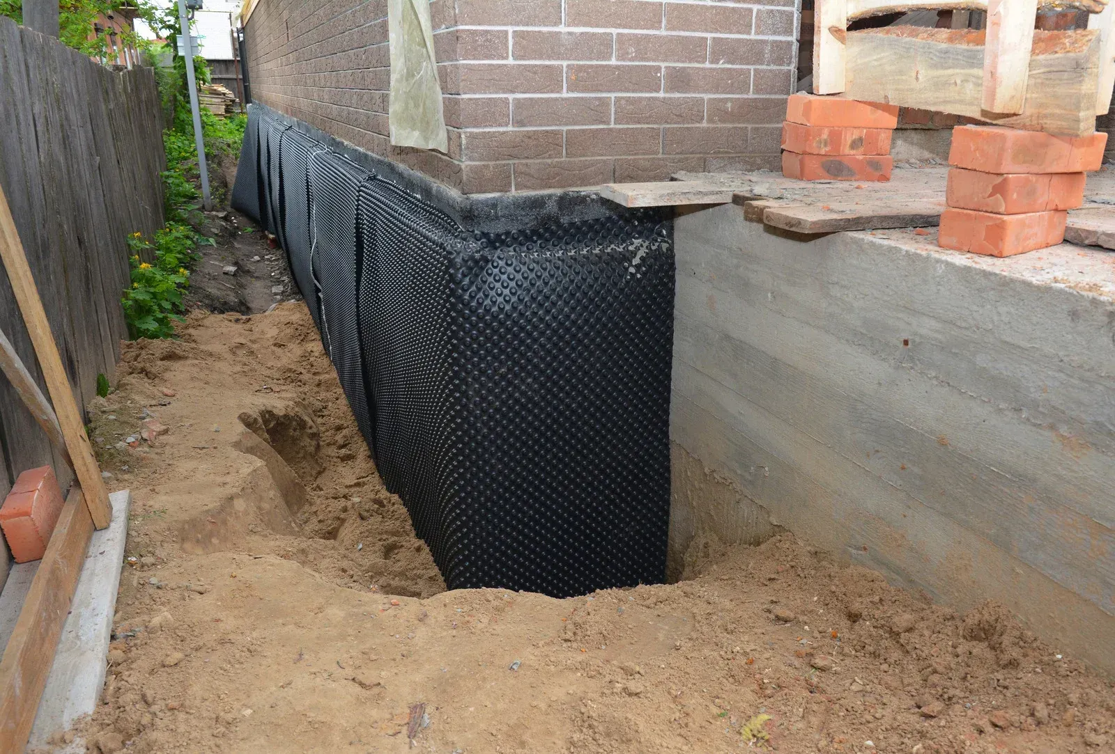Backyard foundation wall wrapped in black waterproofing membrane beside a dirt trench and concrete slab