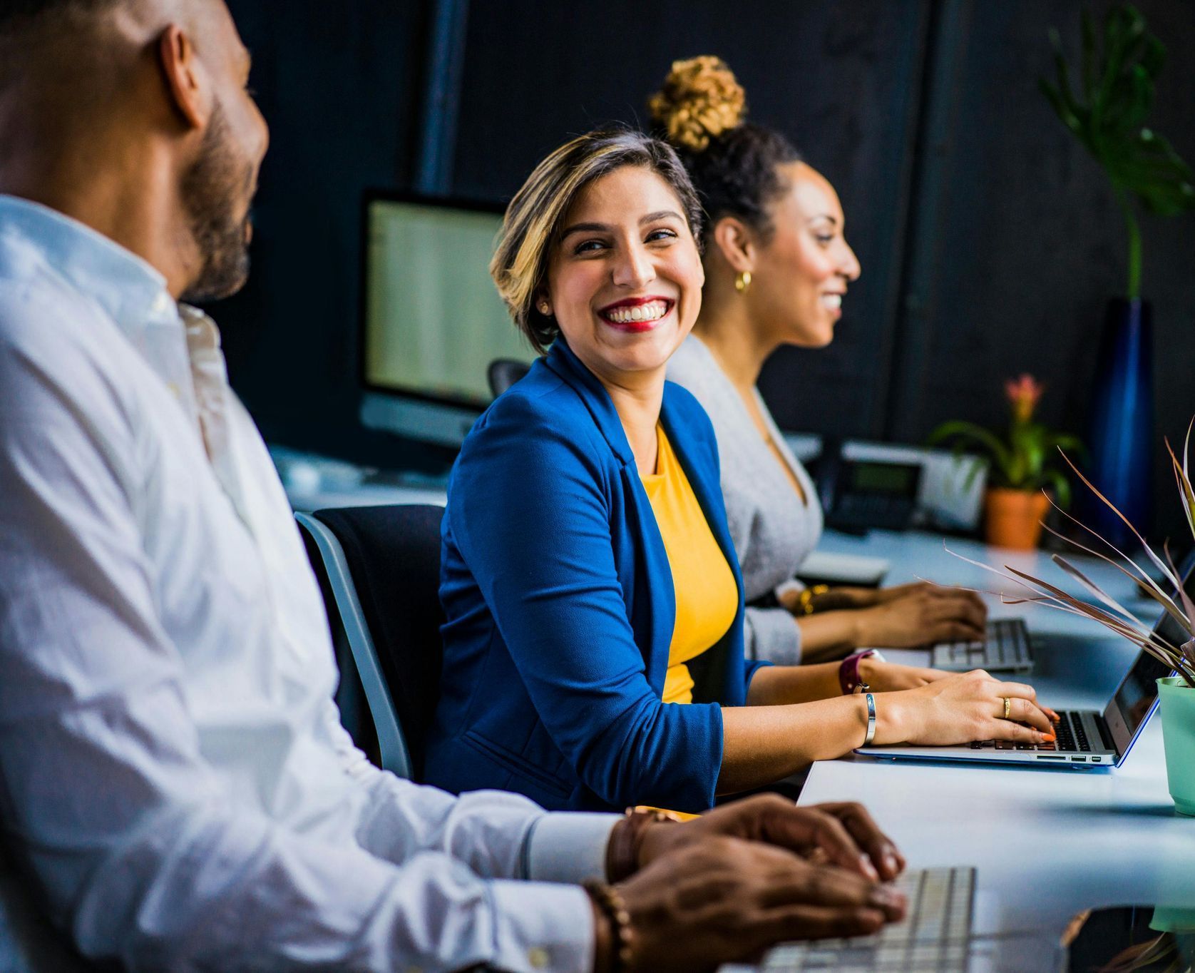 Three diverse colleagues working at computers in an office. The woman in the center smiles at the viewer.