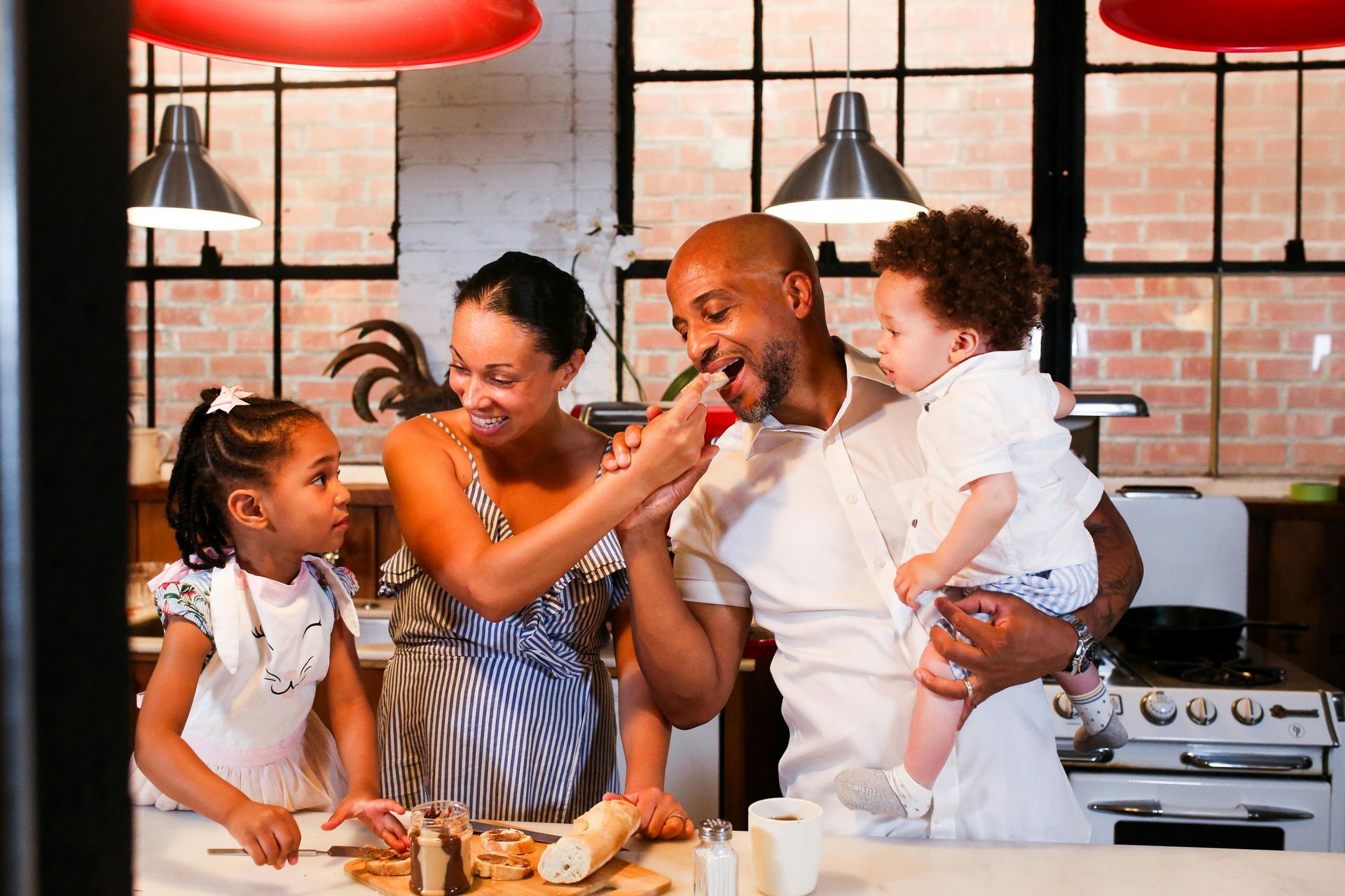 Family in a kitchen: Parents feeding tomato to child, daughter watches. Brick wall, red lights.