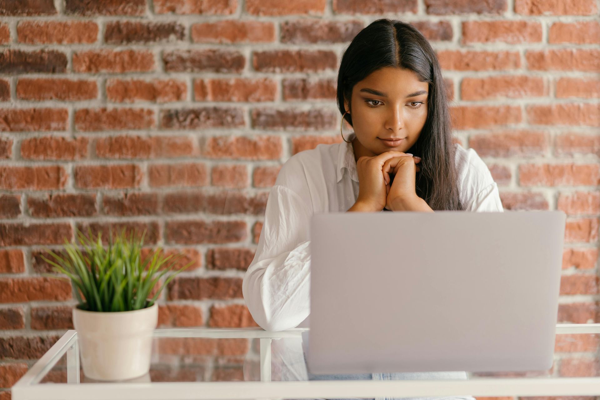 Woman with dark hair looking at a laptop, resting chin on hands. Sitting at a glass table with a brick wall backdrop.