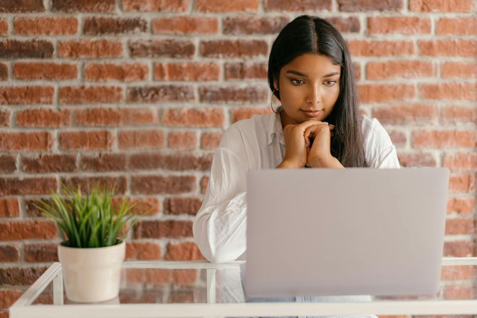 Woman with dark hair looking at a laptop, resting chin on hands. Sitting at a glass table with a brick wall backdrop.