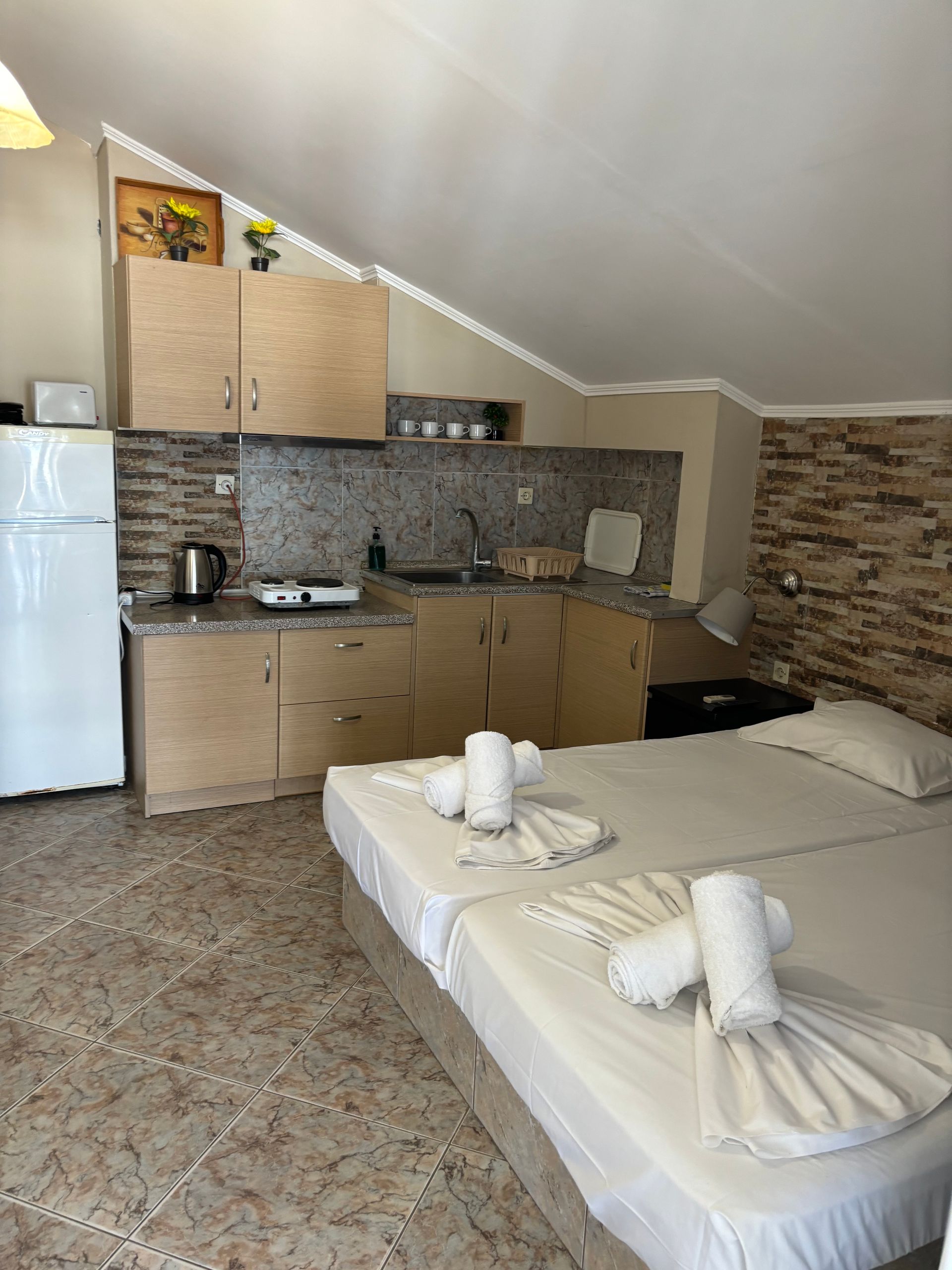 Small kitchen with beige cabinets, tiled backsplash, white fridge, and a bed in the foreground.