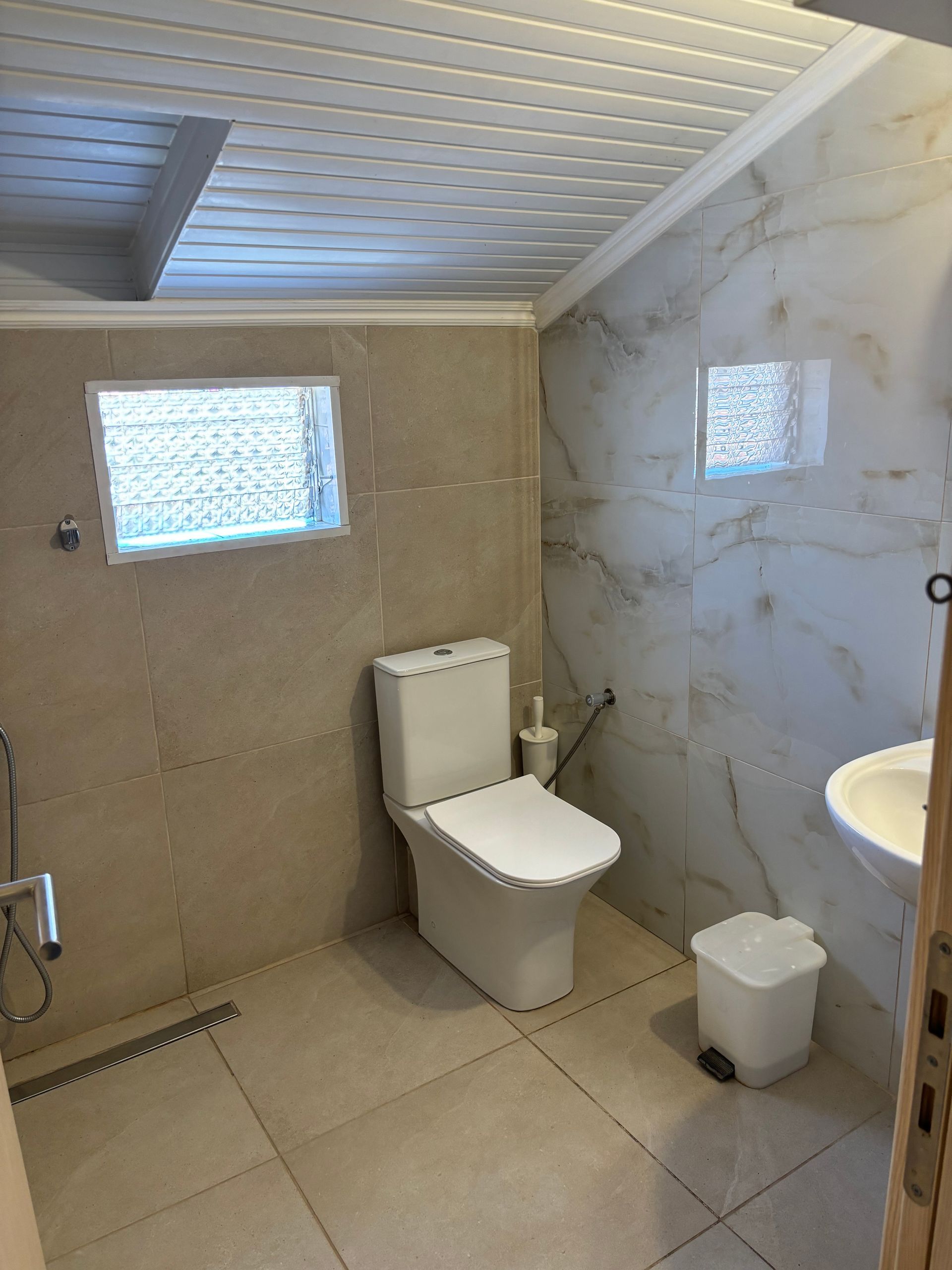 Modern bathroom with beige tile, white toilet, small window, sink, and trash bin