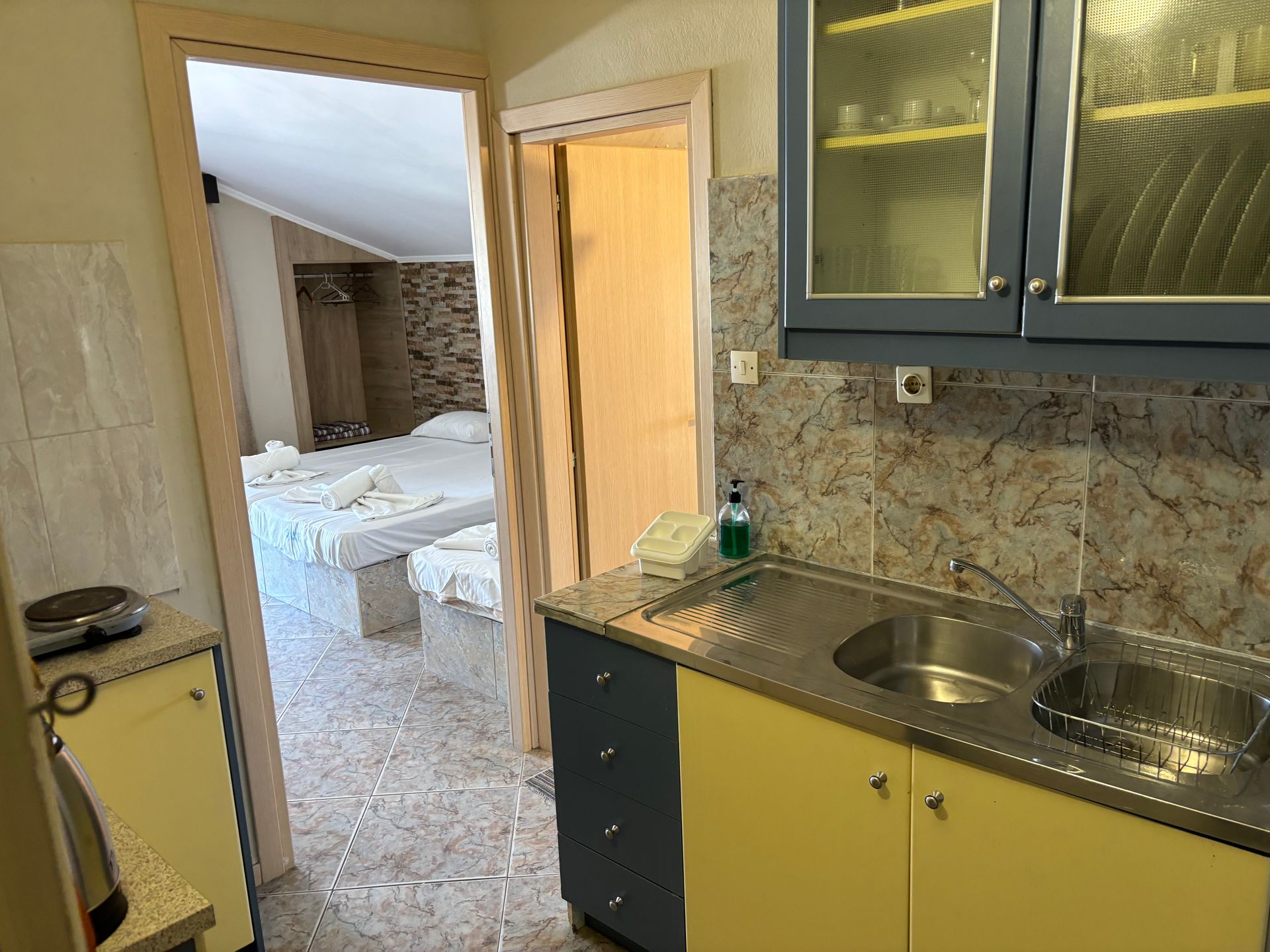 Small kitchen with yellow cabinets and stainless sink, opening to a bedroom through a doorway.