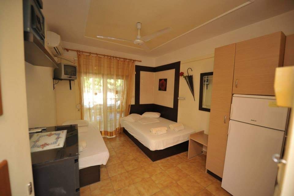 Small hotel room with two single beds, a window with curtains, and a wardrobe beside a mini-fridge.