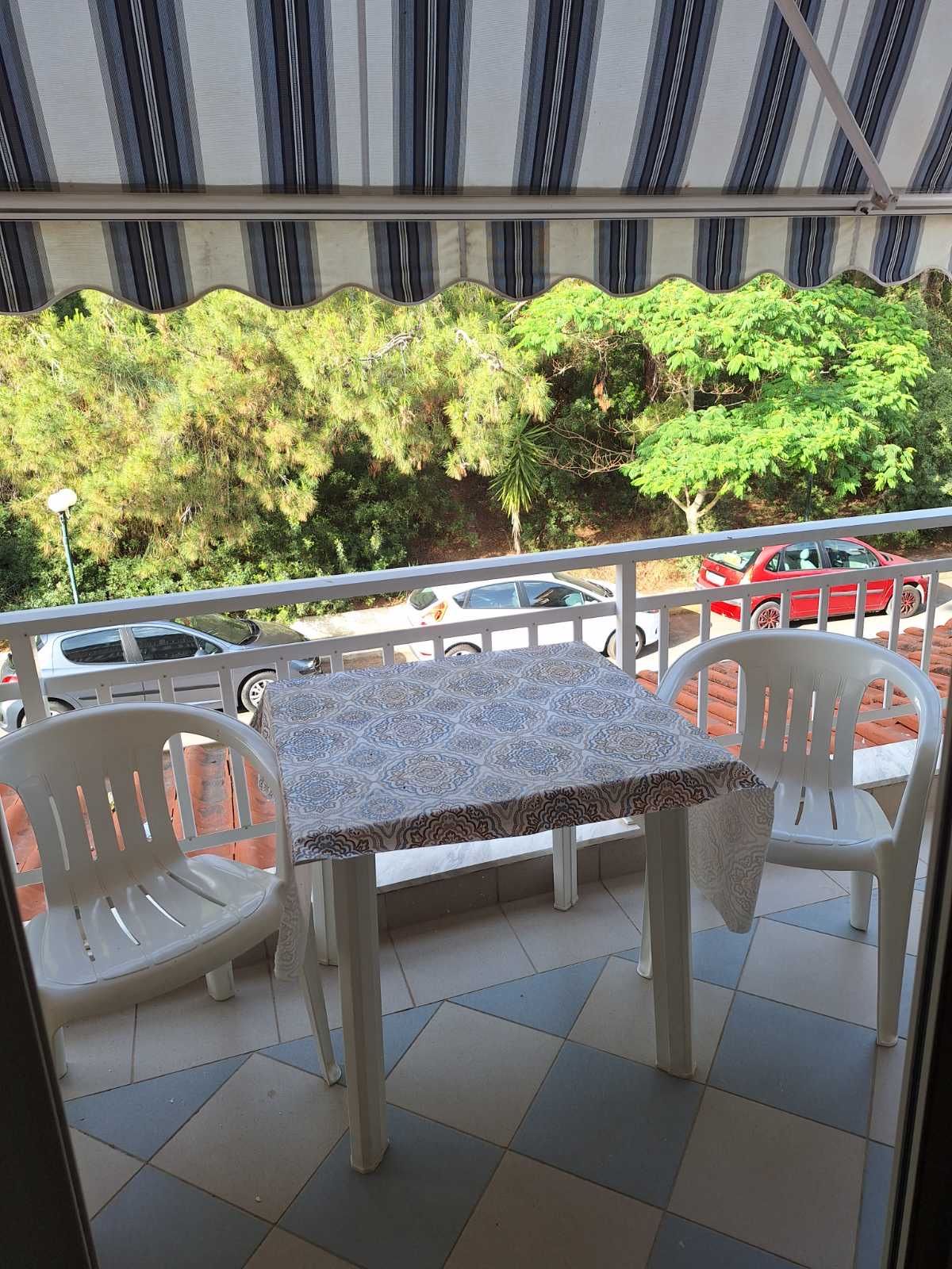 Balcony table with two white chairs under a striped awning, overlooking trees and parked cars.