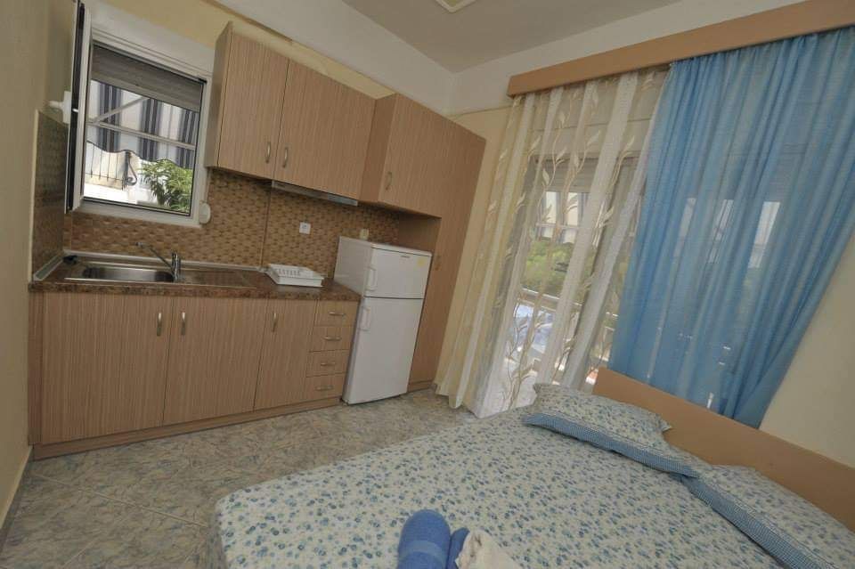 Small bedroom with kitchenette, bed, white fridge, and blue curtains by a sliding door.