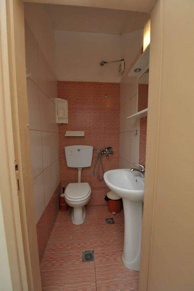 Narrow pink-tiled bathroom with toilet, sink, showerhead, and wall shelves