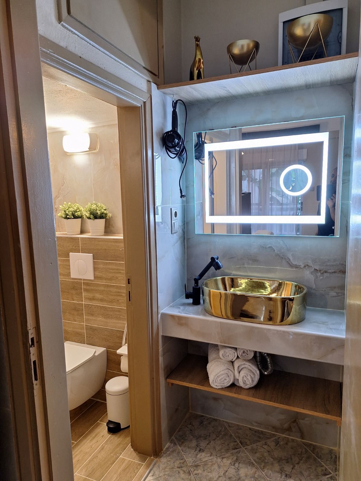 Compact bathroom vanity with lit mirror, gold sink, and open shelves beside a toilet.