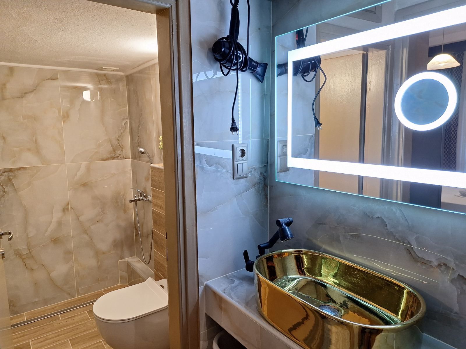 Modern bathroom with marble shower, toilet, and gold sink under a backlit mirror