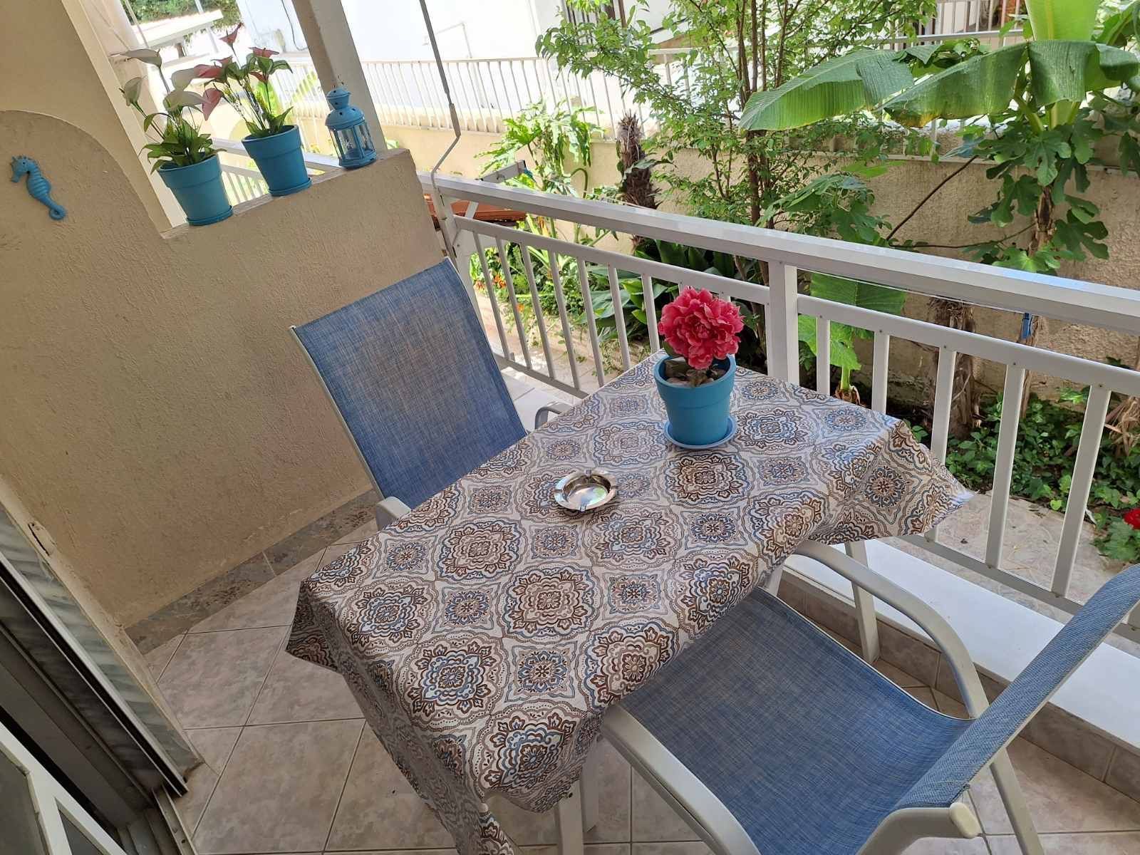 Small patio table with blue chairs, patterned tablecloth, potted red flowers, and a balcony view