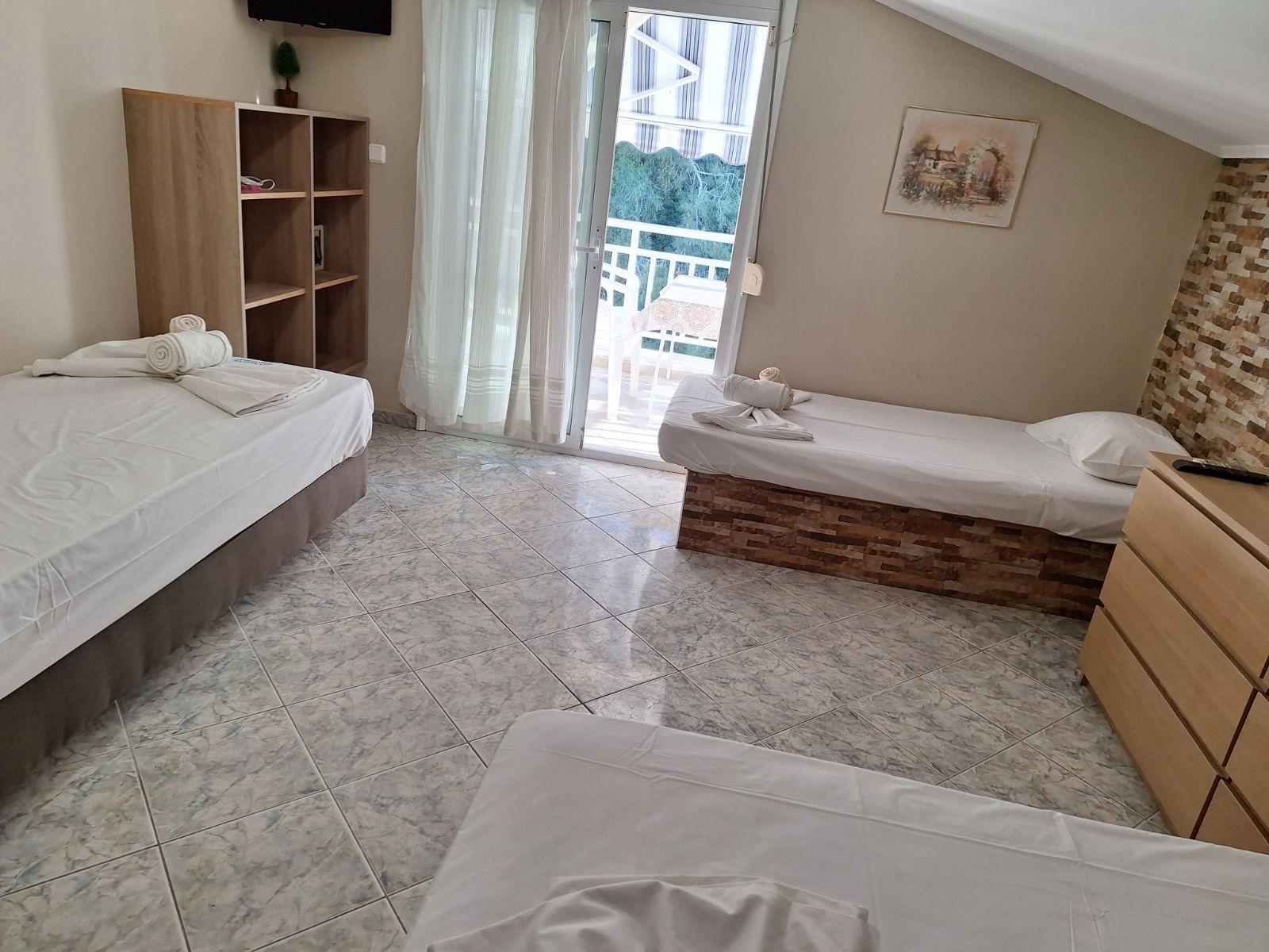 Small bedroom with three single beds, tiled floor, shelves, and a balcony door with sheer curtains.