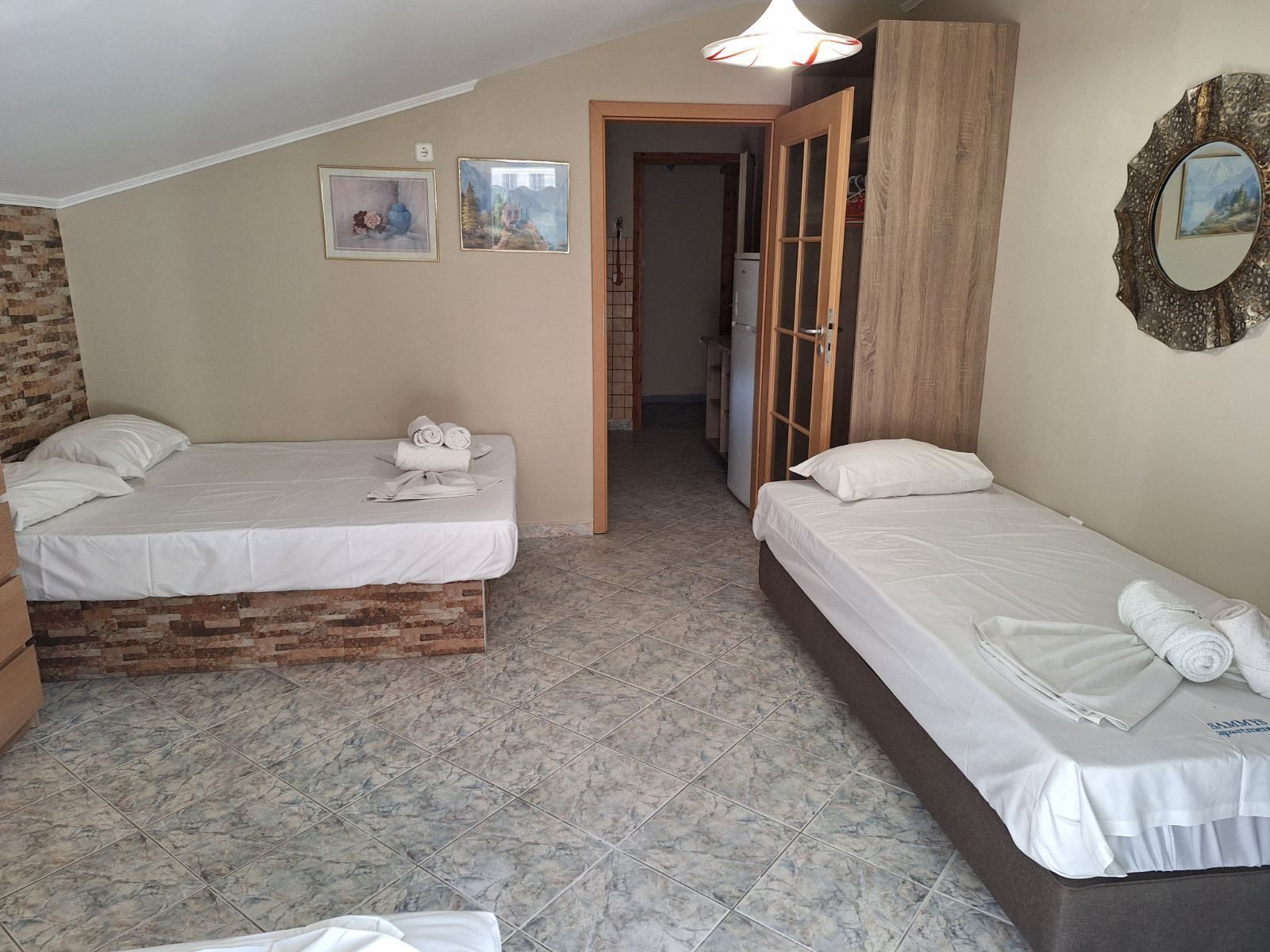Small bedroom with two single beds, tiled floor, beige walls, and an open doorway to a hall.