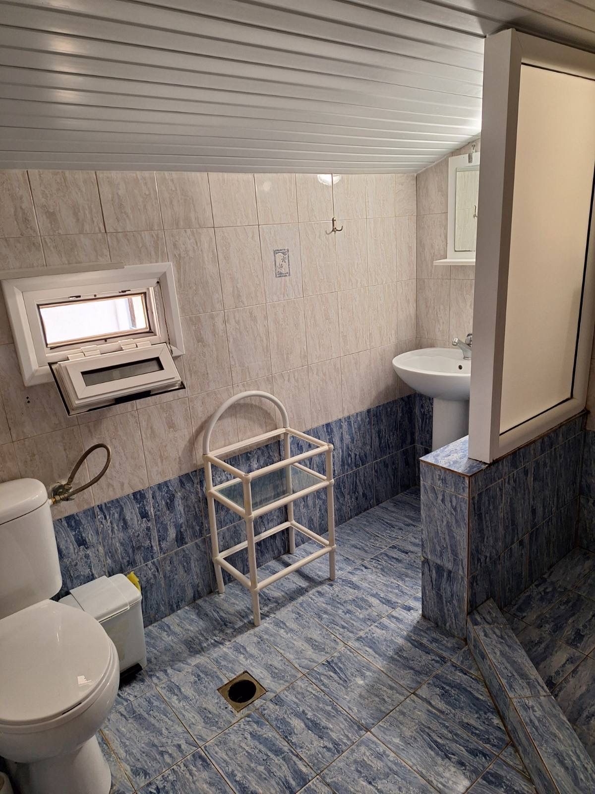 Small tiled bathroom with a toilet, sink, shower, and a wooden shelf in the center.