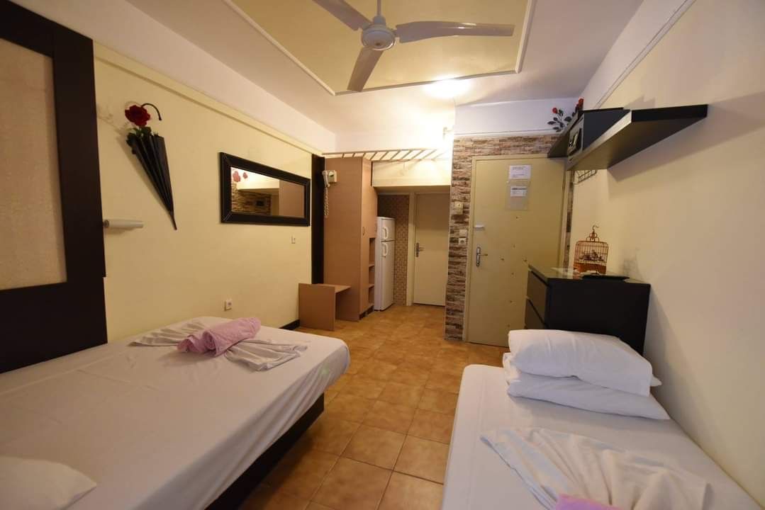 Small hotel room with two single beds, ceiling fan, wall art, and open doorway to a hallway.