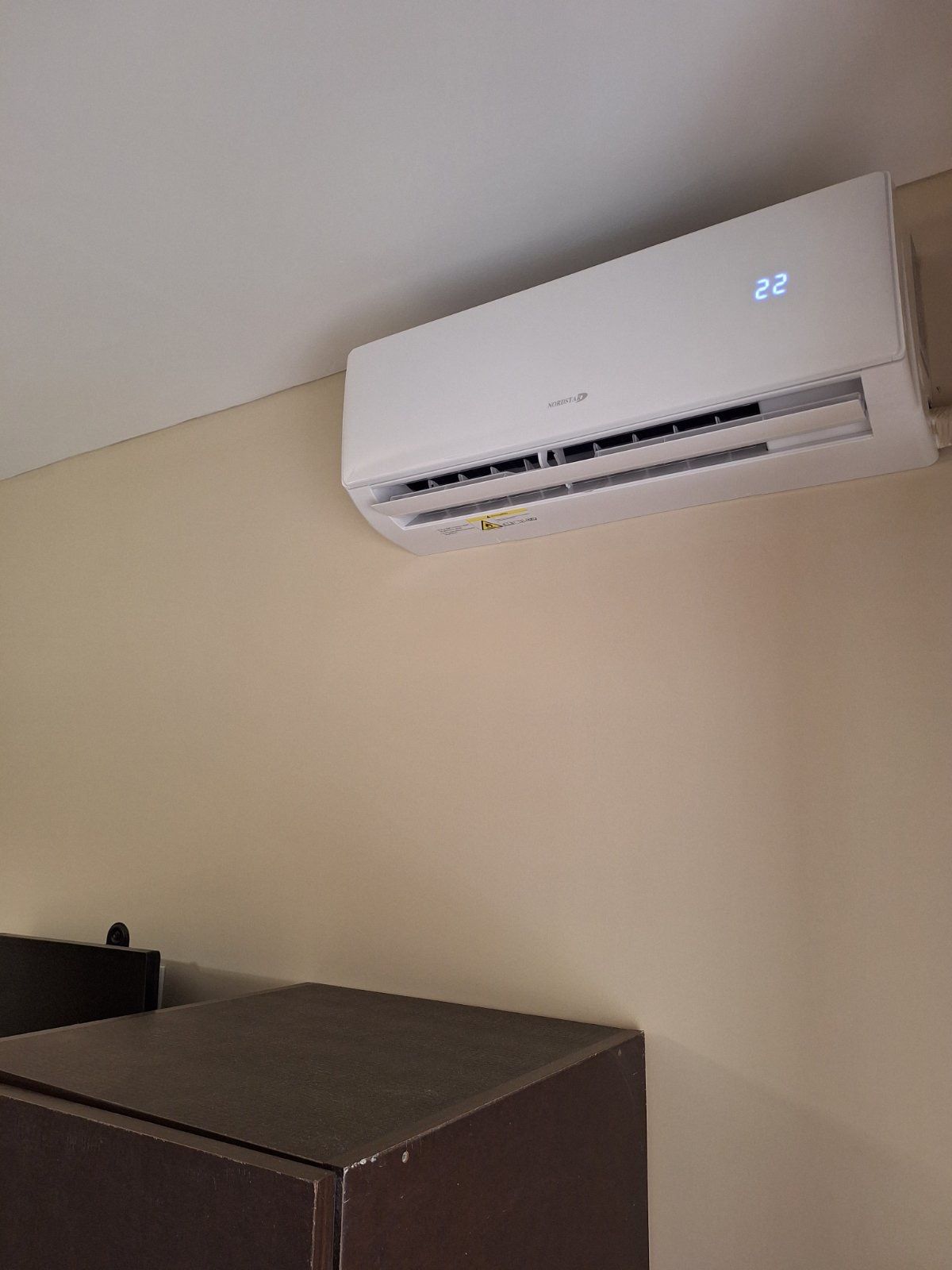 White wall-mounted air conditioner above a dark wooden cabinet in a beige room.