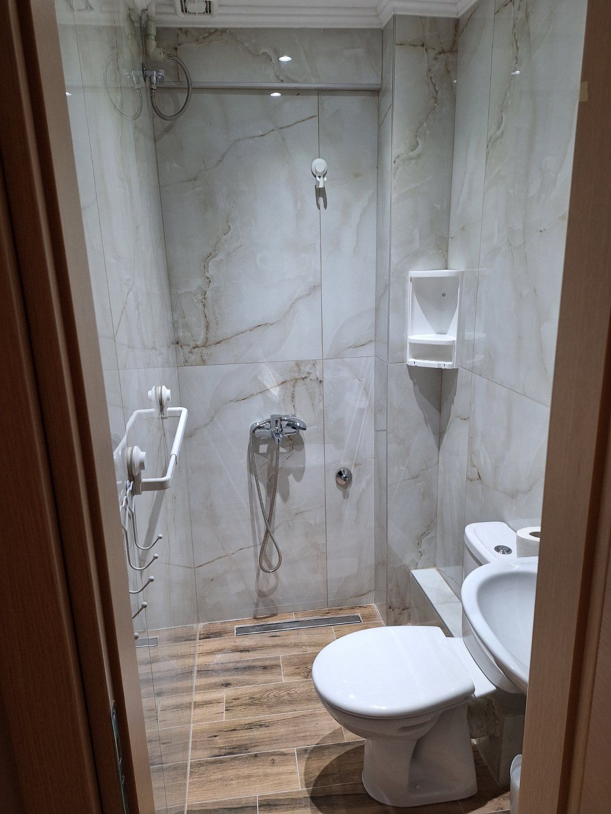 Small marble-tiled bathroom with toilet, wall-mounted sink, grab bar, and shower fixtures.