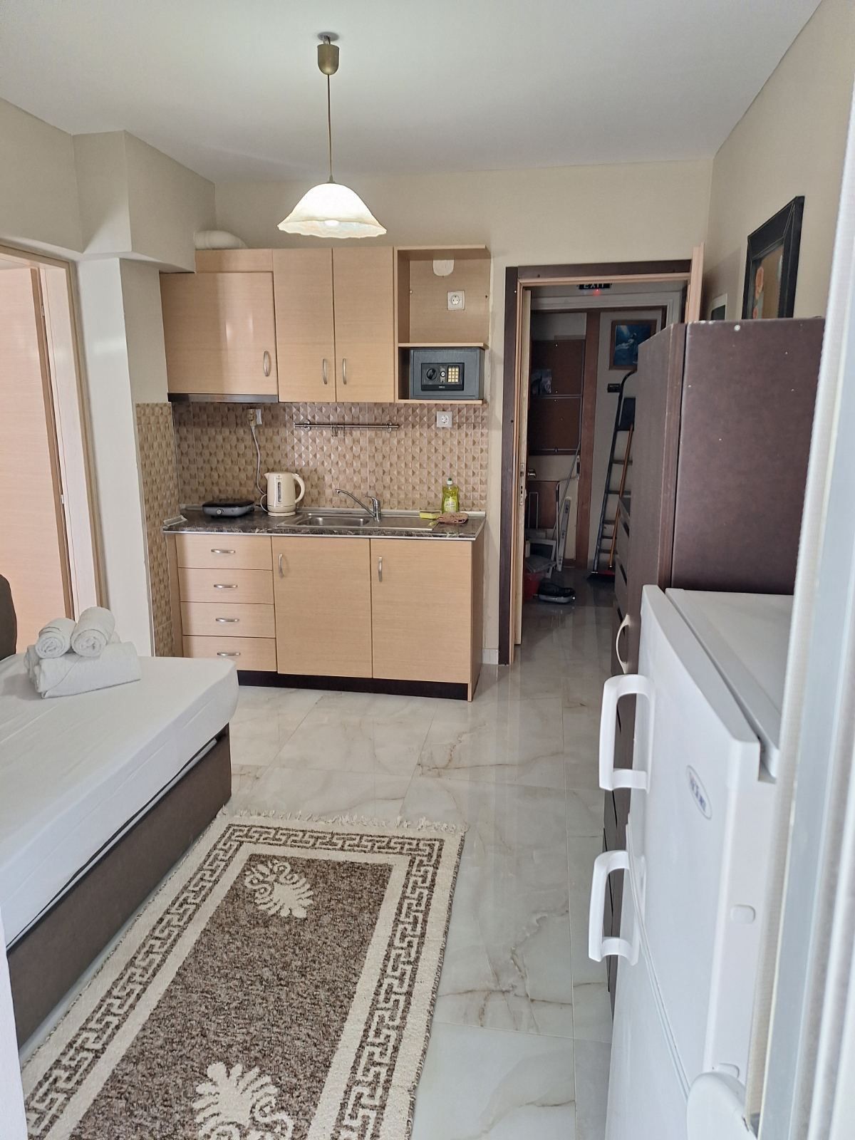 Compact kitchenette with tan cabinets, tiled backsplash, marble floor, white appliances, and a small rug.