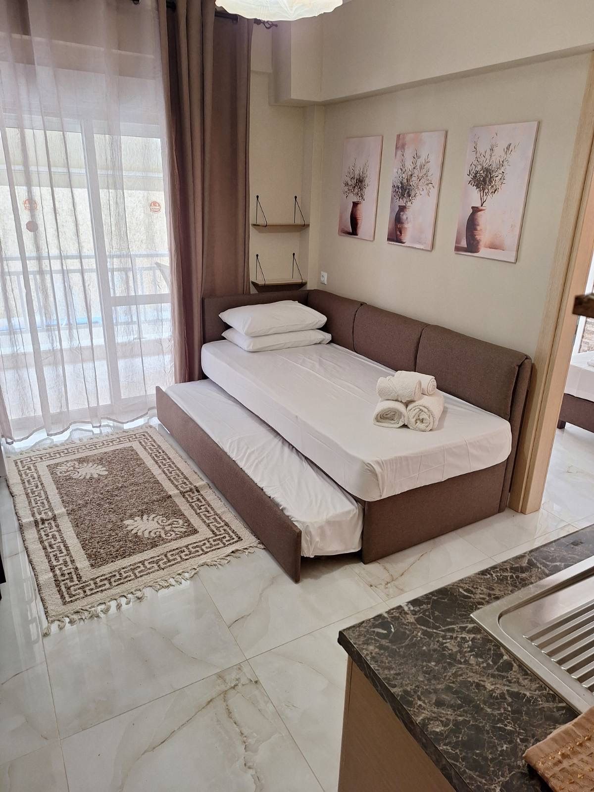 Compact bedroom with a single bed, sheer curtains, wall art, and a patterned rug beside a window.