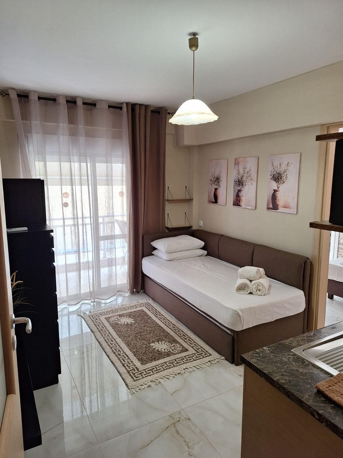 Small bedroom with a single bed, sheer curtains, wall art, rug, and kitchen counter in the foreground.