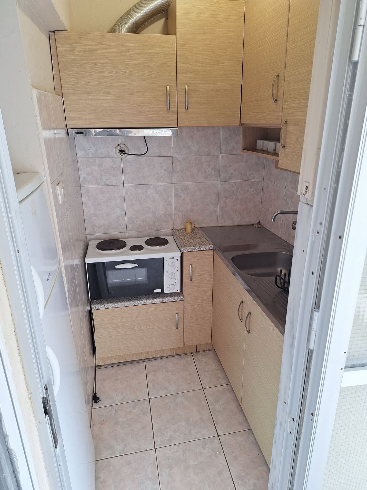 Small beige kitchenette with cabinets, microwave, sink, and tiled floor in a narrow alcove.