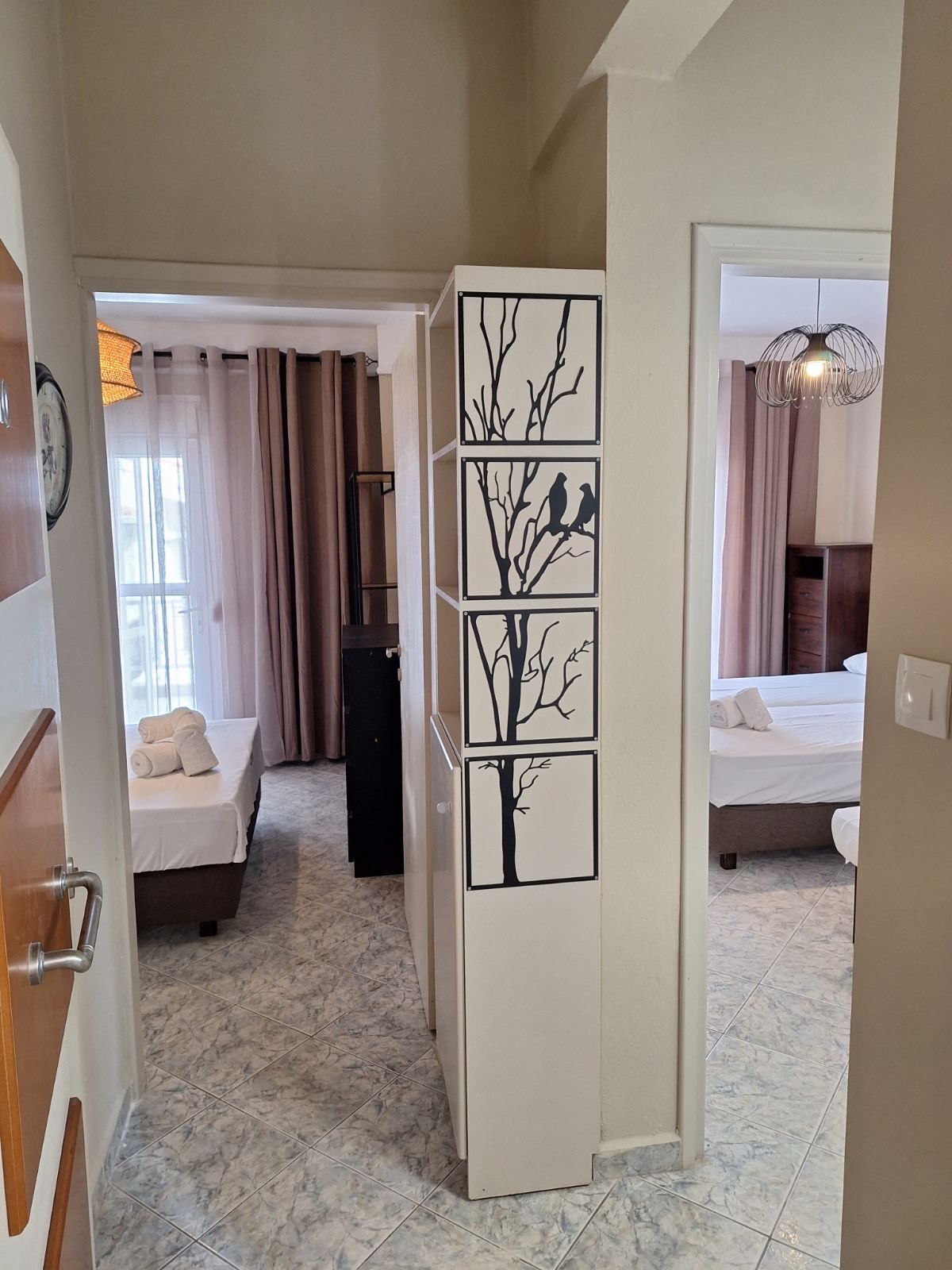 Hallway view into a bedroom with a bed, pink curtains, and framed tree artwork on a wall panel.