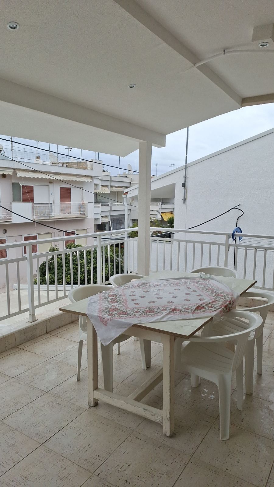 Covered balcony with white plastic table and chairs overlooking neighboring buildings and sky.