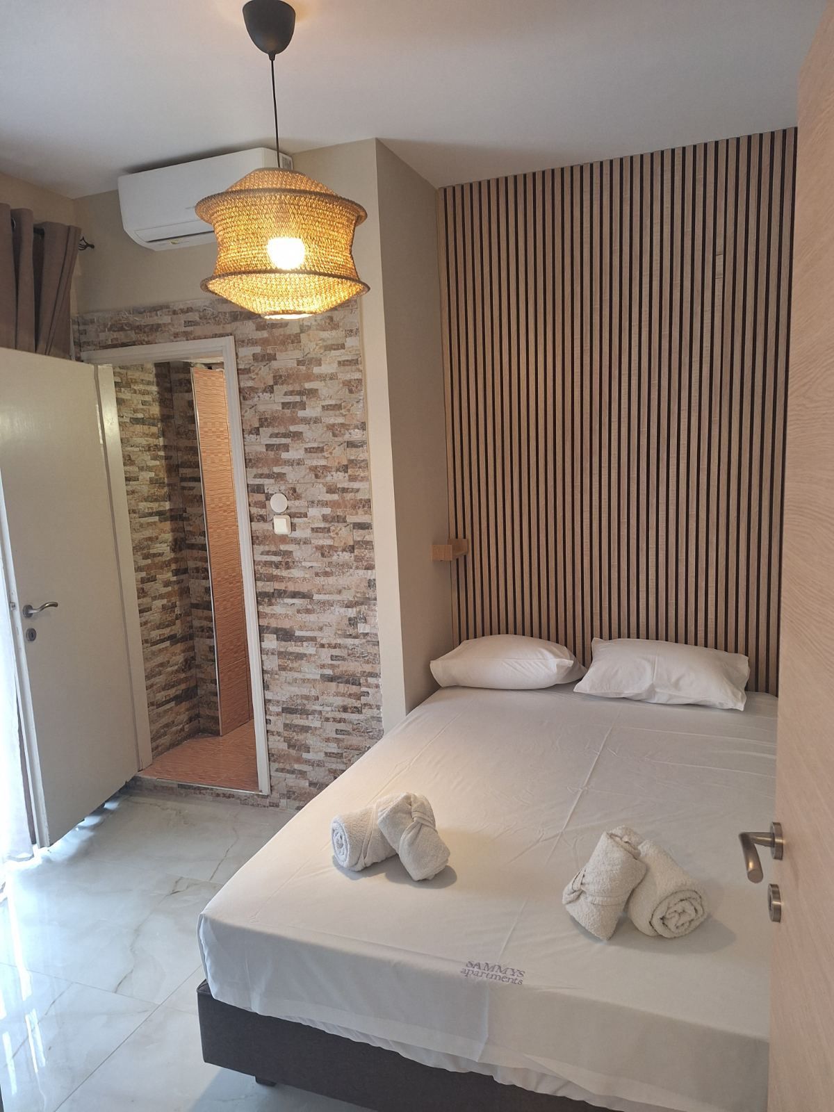 Small bedroom with a neatly made bed, striped accent wall, wicker pendant light, and open door to a tiled area.
