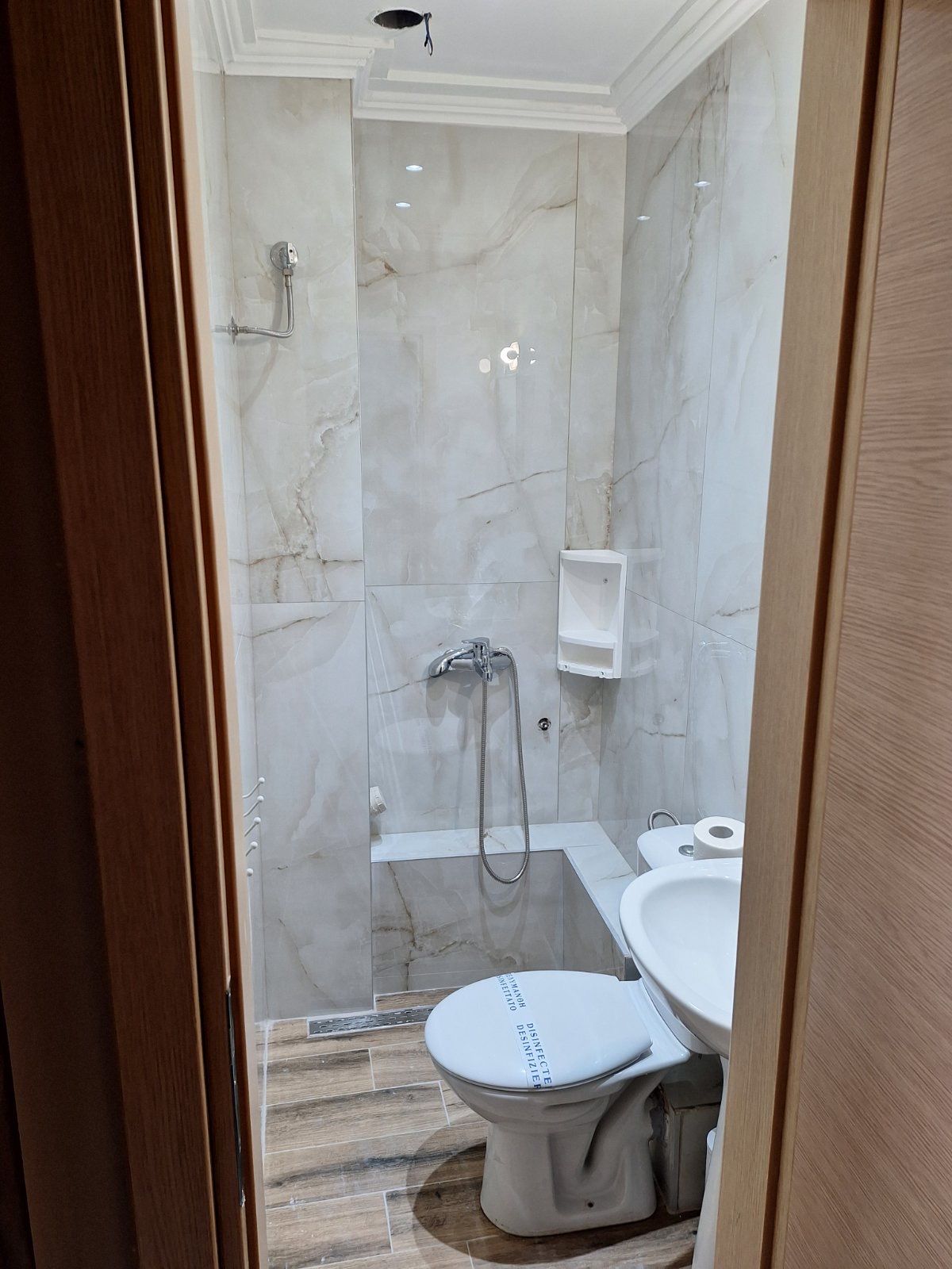 Small tiled bathroom with toilet, shower hose, and marble-pattern walls, viewed through a doorway
