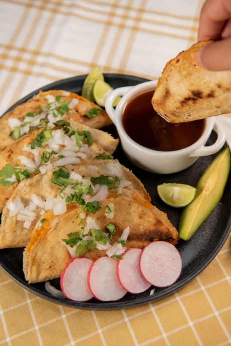 A hand dips a crispy taco into a small bowl of sauce, served on a plate with fresh radish slices, lime, and avocado.