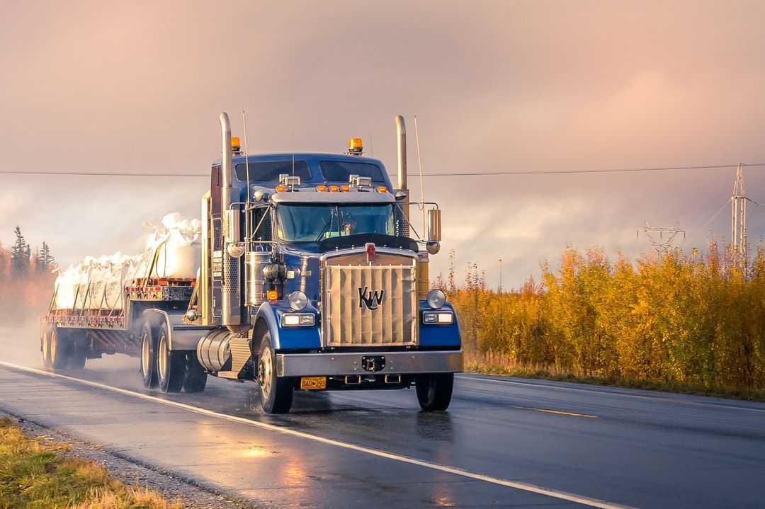 Blue semi-truck driving on a wet road in an industrial area at sunset