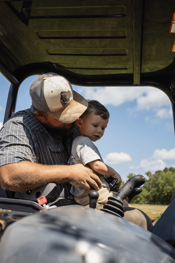 Man wearing cap, teaches young child to operate machinery outdoors.