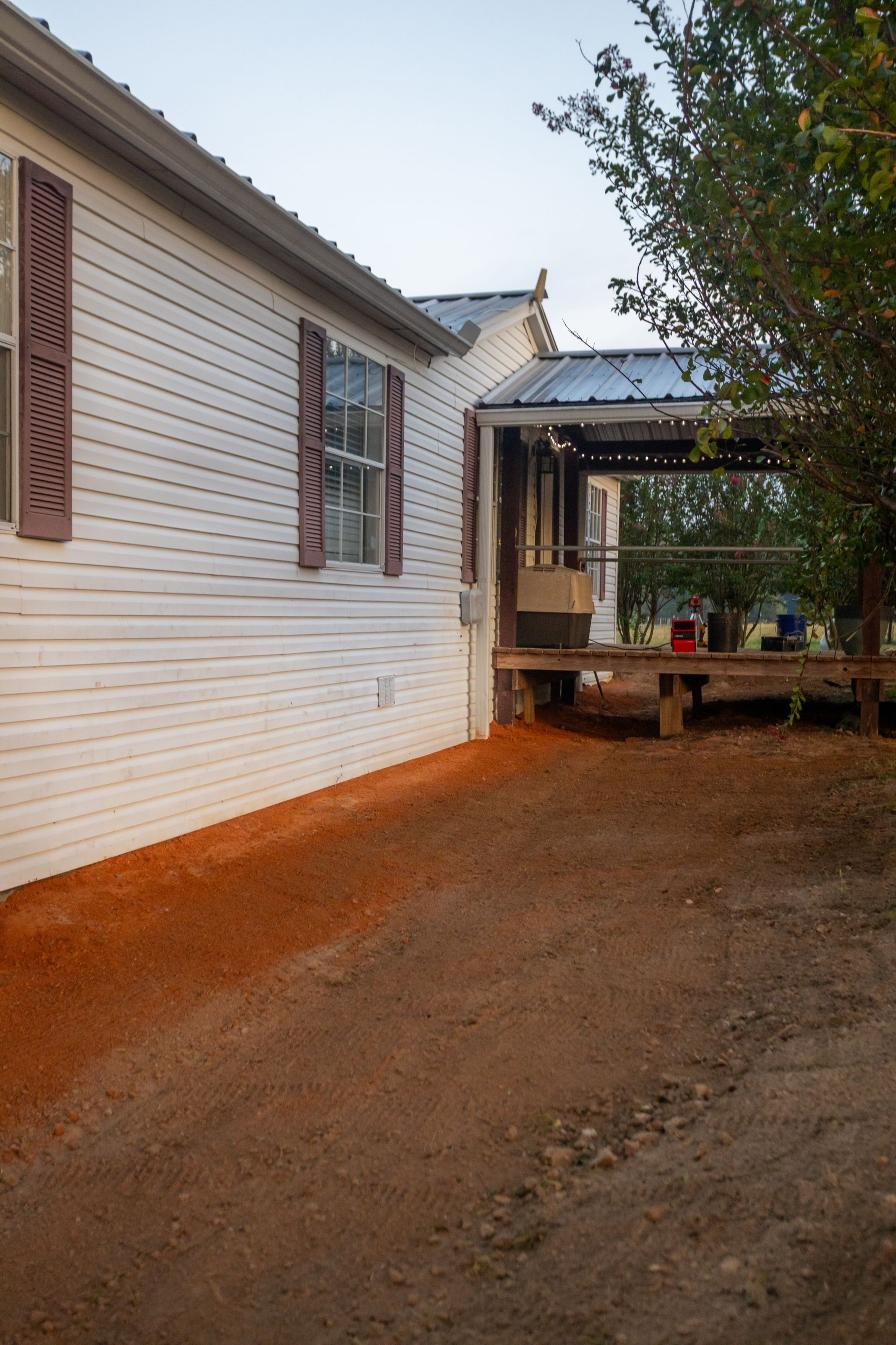 White house with brown shutters, next to a dirt pathway leading to a wooden porch.