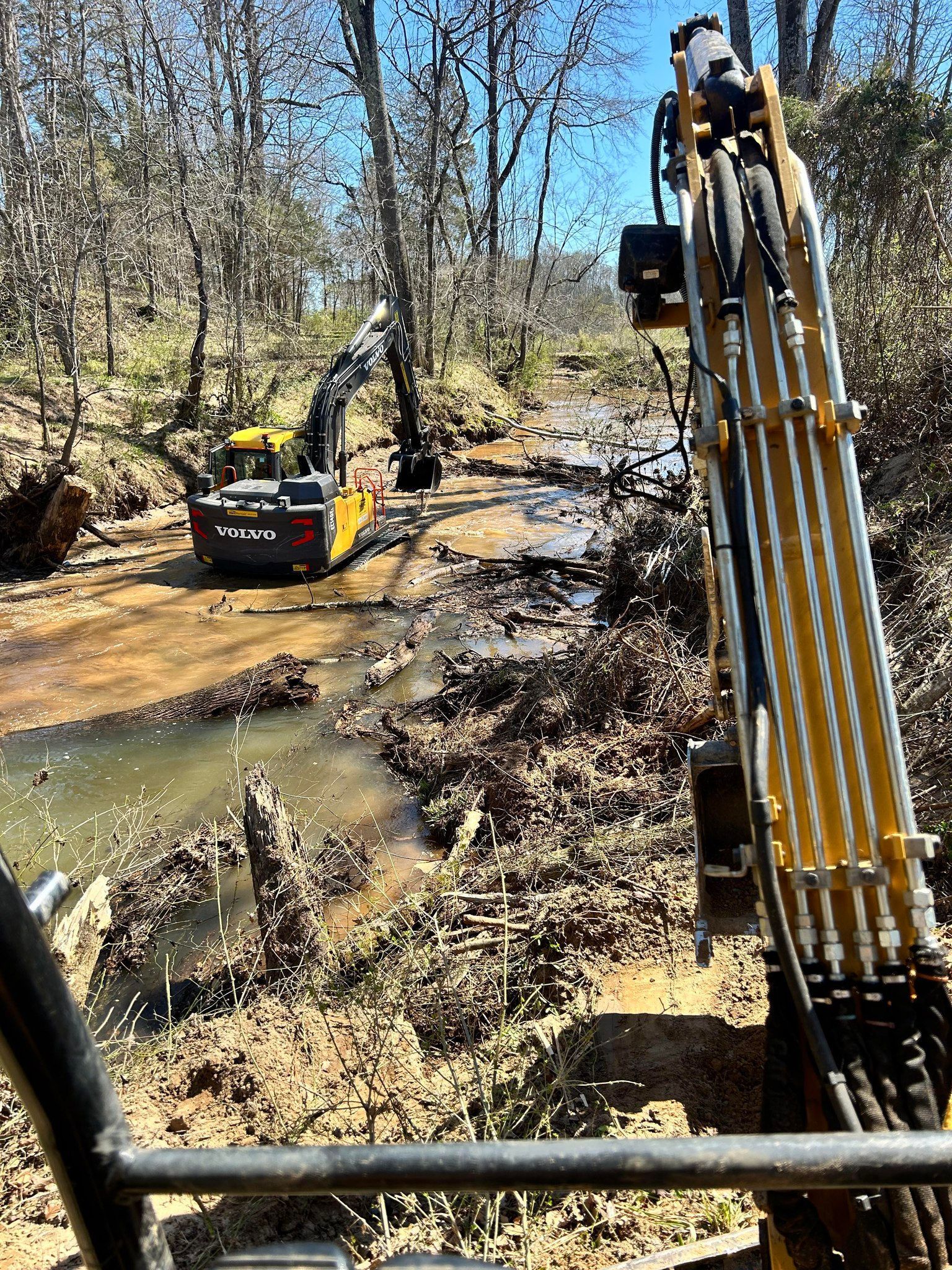 Two yellow excavators clearing a muddy creek, trees in background.