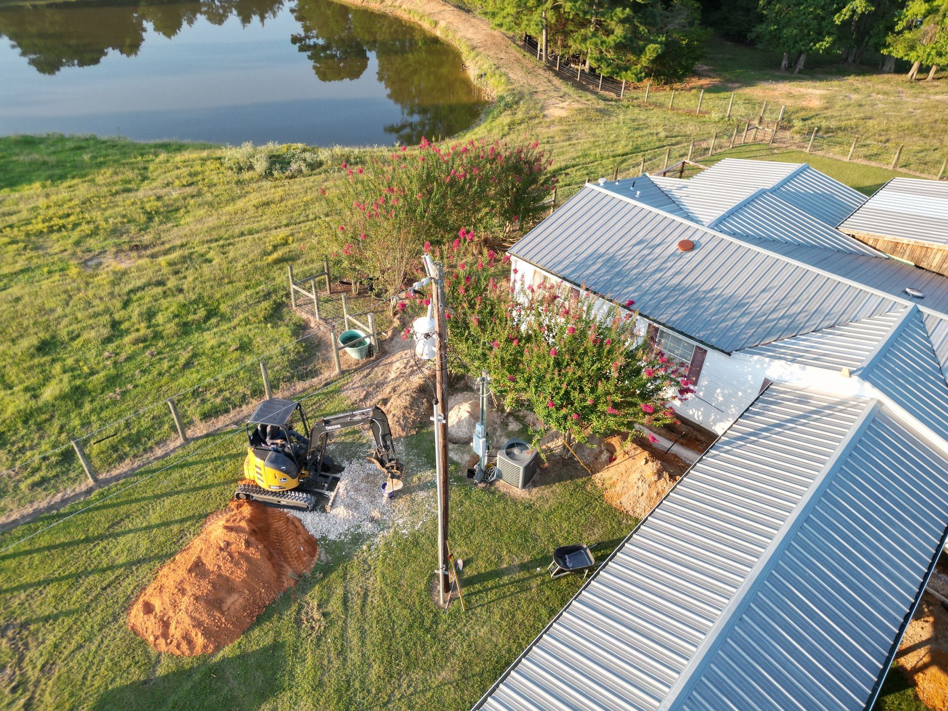 Construction site near a lake with an excavator, a house with a metal roof, and piles of dirt.