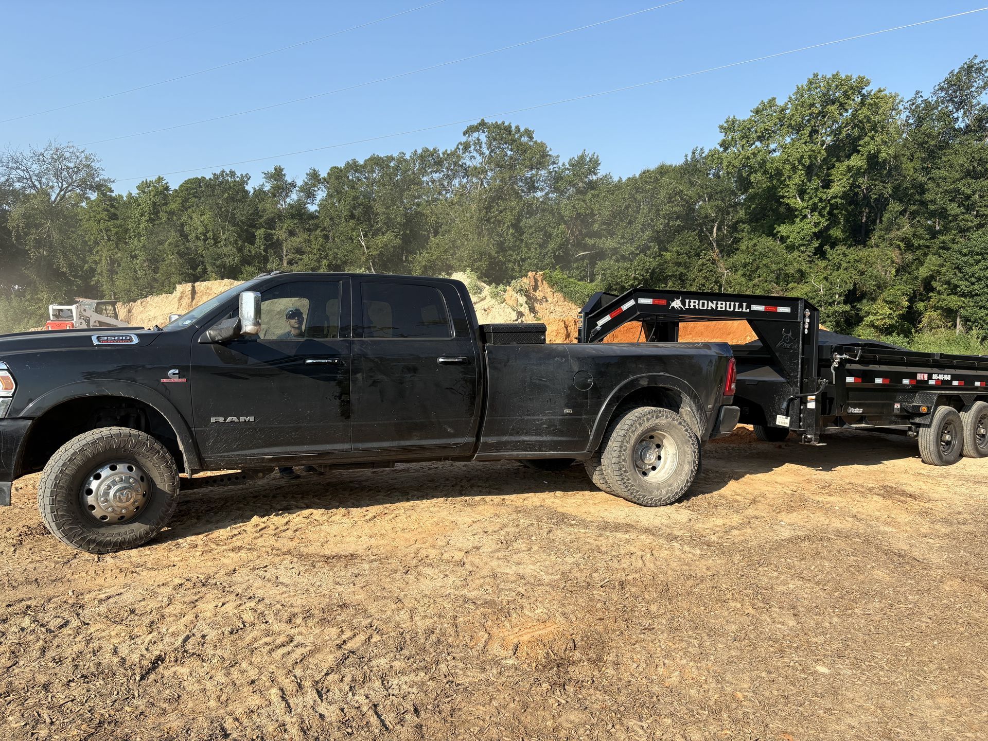 Dark pickup truck towing a black trailer on a dirt surface. Trees and blue sky in the background.