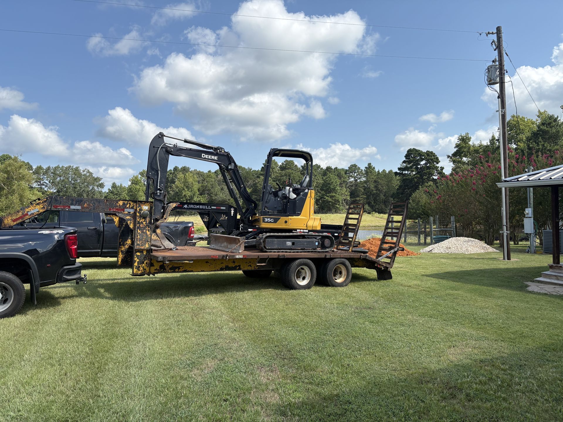 A yellow excavator on a trailer, towed by a black truck, in a grassy yard under a cloudy sky.