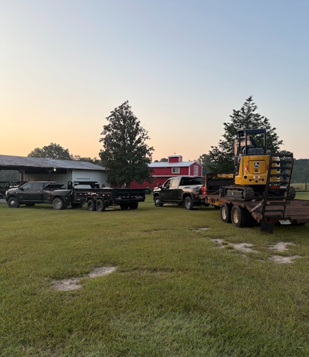Two trucks with trailers on a grassy field, one hauling a yellow construction machine, with a red barn in the background.