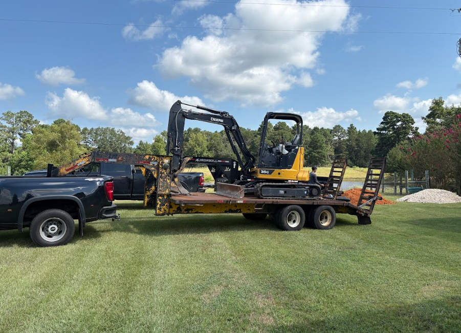 Black pickup truck towing a yellow excavator on a flatbed trailer, parked on grass.