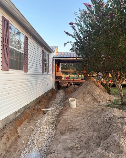 Trench dug along a white house for drainage, with a pile of sand and a porch in the background.